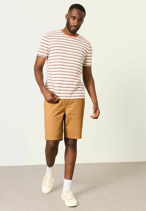 Mann trägt beige Shorts, weiße Sneakers mit Socken und ein weißes Kurzarmhemd mit roten Streifen, steht vor einem einfarbigen hellen Hintergrund.