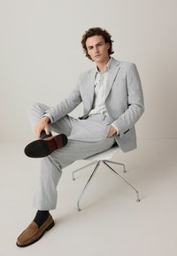 Traje gris claro con tejido texturizado, corte relajado, combinado con una camisa clara de estampado. Mocasines marrones y calcetines oscuros completan el look.