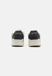 Lacoste sneaker fekete bőr felsőrész, fehér díszítések és bézs gumitalp kombinációjával. Jellemzője a logó a sarokrészen és a sima textúra.