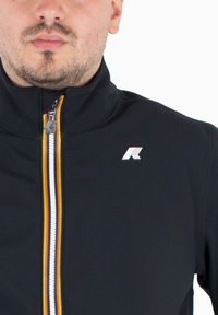 Zwarte ritsjack gemaakt van glad materiaal. Heeft een hoge kraag, witte rits, een veelkleurige streep en een klein logo op de borst.