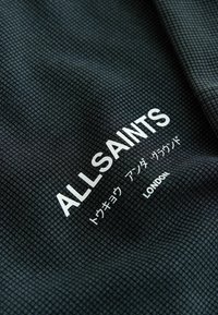 Sort tekstureret stof med hvid tekst, der lyder "ALLSAINTS," japanske tegn og "LONDON" trykt diagonalt.