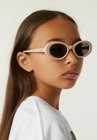 Lunettes de soleil ovales beige clair avec des verres sombres, présentant une monture en plastique lisse et des branches fines. Portées par un enfant avec des cheveux bruns raides.