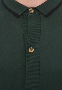 Σκούρο πράσινο πλεκτό πόλο μπλουζάκι με μαύρο γιακά, με ένα μπεζ κουμπί στον λαιμό και ριμπ υφή κατά μήκος της επένδυσης.