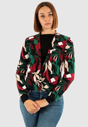 Femme avec des cheveux auburn raides portant un blazer floral multicolore par-dessus un top noir et un jean bleu, se tenant les mains jointes.