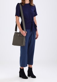 Tmavě modrý krátký rukáv s detailem na zavazování, modré široké kalhoty, šedá kabelka přes rameno a černé kotníkové boty na osobě. Jednoduché pozadí.