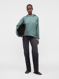 Femme portant un sweat à capuche bleu sarcelle et un jean noir, tenant une veste noire pliée, debout avec une posture détendue et une expression neutre.