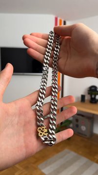 Hopeaketju, jossa on paksut lenkit, jossa on kullanvärinen 'Hugo'-tagi. Suora pinta, kiiltelevä viimeistely, pidetään kädessä sumeaa taustaa vasten.