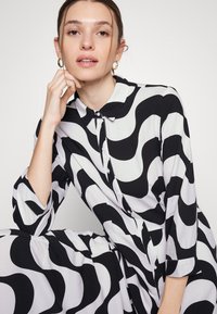 Vestido em padrão ondulado preto e branco com gola, botão na frente e mangas até ao cotovelo. Tecido suave com um acabamento texturizado.