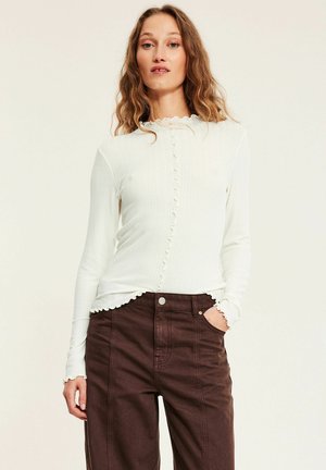 Kvinde med bølget hår iført en hvid ribstrikket langærmet top med flæsekant og mørkebrune bukser med høj talje, hånd i lommen.