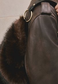 Geantă din piele maro cu finisaj texturat și un accent din blană pufoasă maro închis. Are un inel circular din metal auriu și o curea neagră.