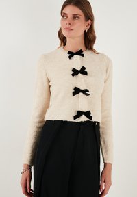 Cardigan beige in maglia pelosa con quattro fiocchi in velluto nero per la chiusura, maniche lunghe e design cropped. I pantaloni sono neri.
