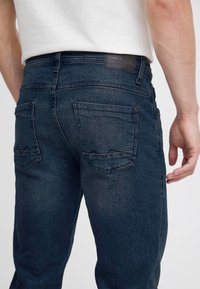 Mørkeblå denimjeans med en lett falmet tekstur. Har to baklommer med et trekantet design og en kontrasterende svart midjebåndslabel.