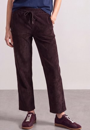 Personne portant un pantalon en velours côtelé marron foncé avec une taille à cordon et des baskets violet foncé avec des lacets blancs, debout sur un sol gris.