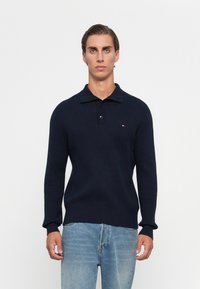 Man met kort, donker haar, gekleed in een marineblauw geribd sweatshirt met twee knopen en lichtblauwe jeans tegen een effen witte achtergrond.