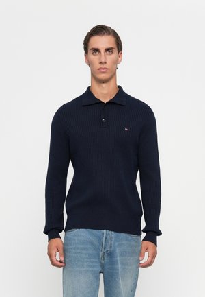 Hombre con cabello oscuro corto que lleva un suéter de punto en color navy con dos botones y unos jeans de color azul claro, frente a un fondo blanco liso.