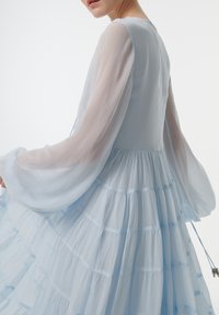 Robe maxi tierée bleu clair avec manches longues transparentes, un corsage ajusté et des couches froncées. Présente un délicat détail en ruban à la taille.