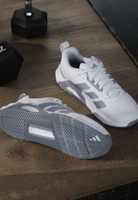 Paire de chaussures de sport blanches et grises sur une surface en bois, une chaussure montrant sa semelle et des haltères noirs en arrière-plan.