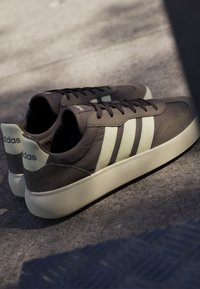 Bruine Adidas-sneakers met witte zolen en drie witte strepen aan de zijkanten, geplaatst op een grijs oppervlak in zacht licht.