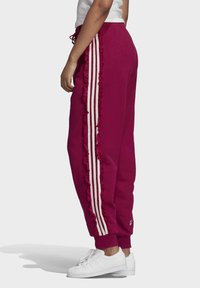 Fuchsiové tepláky s bílými volánkovými pruhy po stranách, zúženými manžetami a šňůrkou v pase. V kombinaci s bílými teniskami.