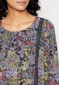 Top com padrão floral e decote redondo, apresentando cores vibrantes, incluindo vermelho, roxo e verde, e um contraste de acabamento escuro ao longo da costura.