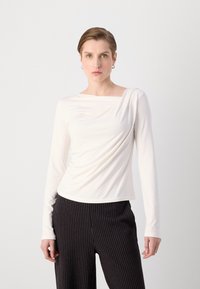 Dlhý rukáv, jemne off-white top s okrúhlym výstrihom a zhromaždenou textúrou. V kombinácii s čiernymi nohavicami s prúžkami pre kontrast.