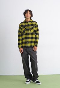 Grøn- og sortternet flannel skjorte, to brystlommer, knap-front; parret med mørke jeans og sorte sneakers på et grågrønt gulv.
