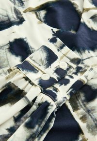 Voogav blouse, millel on abstraktne tumesinine ja oliivroheline muster kreemivalgel taustal, nööbiga detail sääres ja volditud tekstuur.