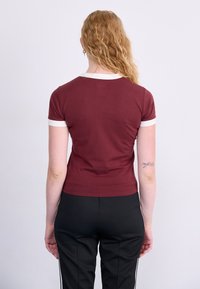 Bordeauxrotes, enges T-Shirt mit weißem Rundhalsausschnitt und Ärmelauschnittern, aus glattem Stoff; kombiniert mit schwarzen Hosen mit weißen Seitenstreifen.