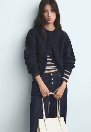 Marine gebreide cardigan met ronde halslijn en knoopsluiting, gedragen over een bruin-wit gestreepte top, gecombineerd met denim broek en een crèmekleurige handtas.