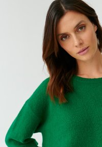 Groene gebreide trui met een ontspannen pasvorm, geribbelde halslijn en textuurafwerking. Model poseert tegen een neutrale achtergrond, met de schouder iets opgeheven.