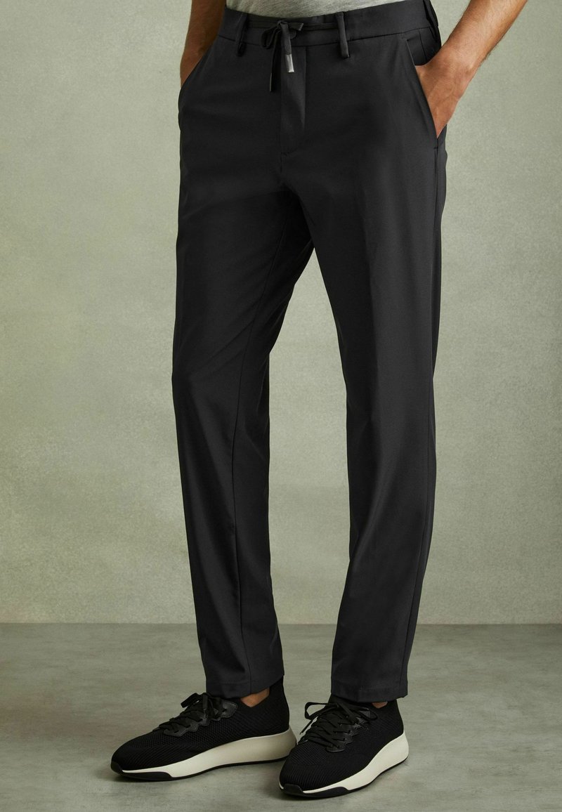Pantaloni negri tapered dintr-un material fin, cu buzunare și o talie cu șnur, asortați cu pantofi sport negri.