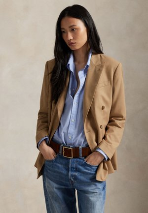 Vrouw met lang donker haar die een beige blazer draagt, een lichtblauw overhemd met knopen, een bruine riem en blauwe spijkerbroek met handen in de zakken.