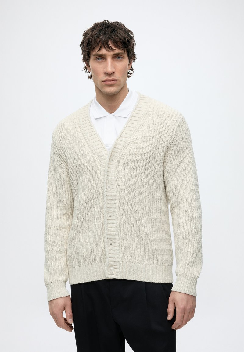 Homme aux cheveux courts bouclés portant un cardigan en maille crème boutonné sur un polo blanc, debout devant un fond clair uni.
