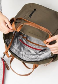 Olijfgroene rugzak met bruine leren banden; interieur heeft een grijs ruitpatroon, een rode ritszak en aanvullende organisatiebanden.