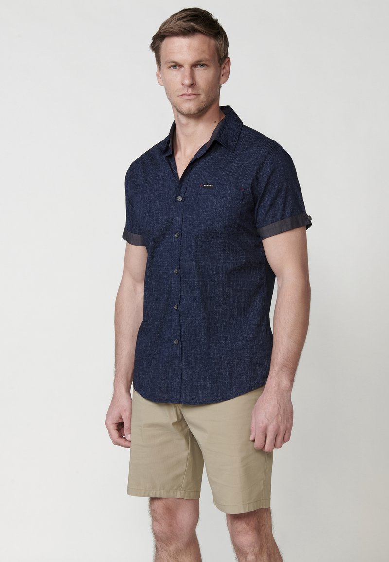 Marineblauw, kortärmige knoopshirt van textuurstof met contrasterende manchetten, gecombineerd met beige shorts. Subtiel logo op de borst.