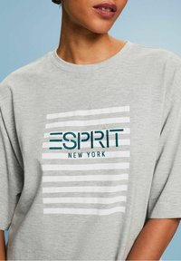 Grå T-shirt med kort ärm, försedd med vita horisontella ränder och broderad text "ESPRIT NEW YORK" i teal. Mjuk textur.