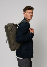 Un sac à dos vert olive en tissu texturé, avec des bretelles rembourrées ajustables et une forme cylindrique élégante, porté par une personne en veste bleue.