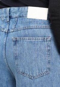 Gros plan d’un jean en denim bleu clair avec une poche arrière et une étiquette blanche vierge, porté avec un haut noir.