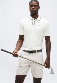Άνδρας που φορά λευκό πόλο μπλουζάκι και σορτς κρατώντας οριζόντια ένα μπαστούνι του γκολφ μπροστά του, πάνω σε ένα απλό φόντο.
