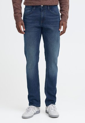 Mann trägt dunkelblaue Jeans, braunen Pullover und weiße Sneaker, steht vor einem einfarbigen hellen Hintergrund.
