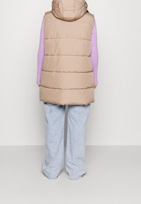 Beige puffer vest med hætte, har vandret syninger. Bæres over en lilla langærmet top og parres med lyseblå brede bukser.