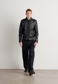 Black leather motorcycle jacket with epaulets, zippered front, and belted waist, paired with loose black pants and black shoes.