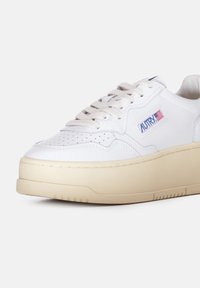 Witte leren sneaker met een chunky beige zool, perforaties op de neus en vetersluiting. Bevat een "AUTRY" logo met een kleine Amerikaanse vlag.