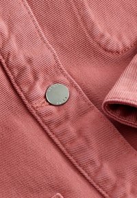 Gros plan sur un tissu denim rose avec un bouton argenté gravé du mot "NEXT" sur un détail de vêtement cousu.