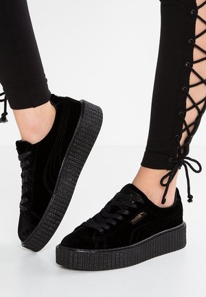 Czarne aksamitne sneakersy na platformie z grubymi, teksturowanymi podeszwami, noszone z czarnymi legginsami z przodu z wiązaniami po bokach.