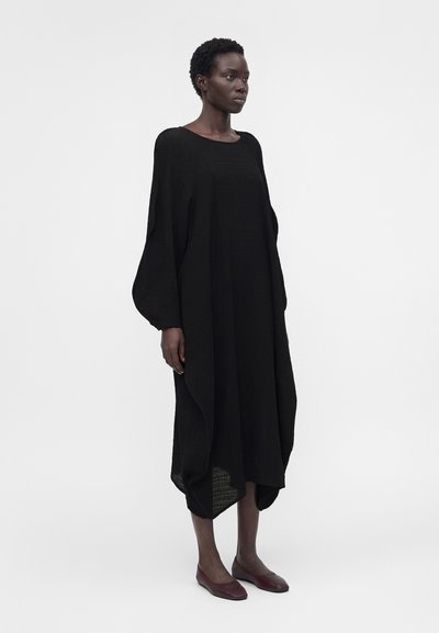 Γυναίκα με κοντά κατσαρά μαλλιά, που φοράει ένα άνετο μαύρο φόρεμα με μακριά μανίκια και σκούρα κόκκινα φλατ παπούτσια, στέκεται μπροστά σε ένα απλό φωτεινό φόντο.