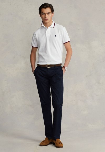 Polo branca com colarinho, calças azul-marinho e mocassins castanhos. O polo tem acabamento vermelho e azul no colarinho, com um logótipo no peito.