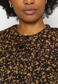 Blusa floral preta com padrões de flores amarelas e vermelhas, gola plissada e fecho de botão, confeccionada em tecido leve.