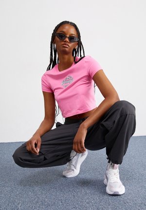 Jeune femme portant des lunettes de soleil noires, un t-shirt court rose, un pantalon cargo noir et des baskets blanches accroupie sur un sol recouvert de moquette.