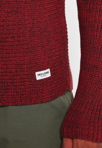 Pusero neulottu syvässä punaisessa, jossa on tekstuuroitu kuvio, varustettuna ribatuilla hihansuilla ja näkyvällä merkkilapulla helman kohdalla.